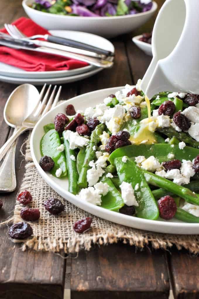 Dressing being poured over a Christmas Salad made with snow peas, asparagus, feta and cranberries.
