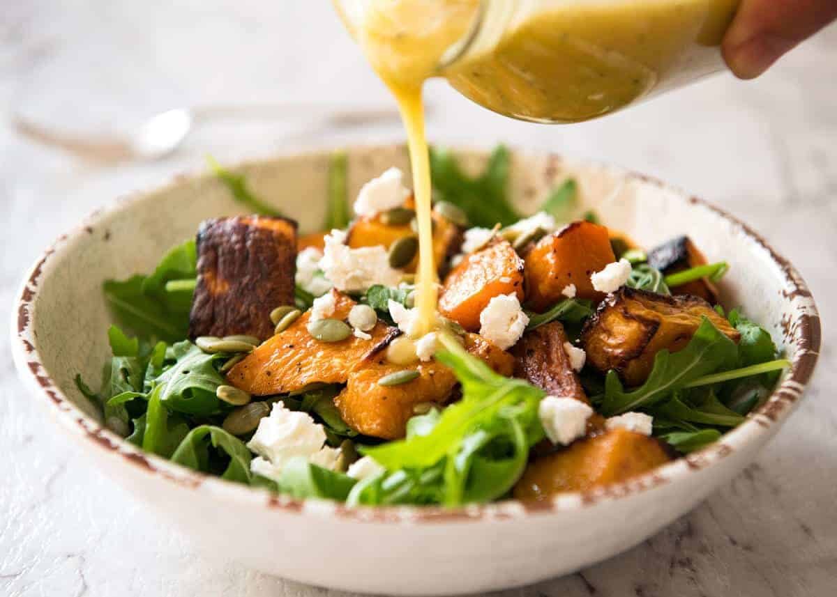 Maple Syrup Dressing being poured over a roasted pumpkin and rocket / arugula salad scattered with feta and pumpkin seeds.