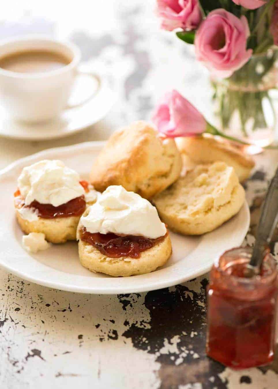 Photo of plain scones on a small white plate with one split open, piled high with jam and cream, ready to be eaten.